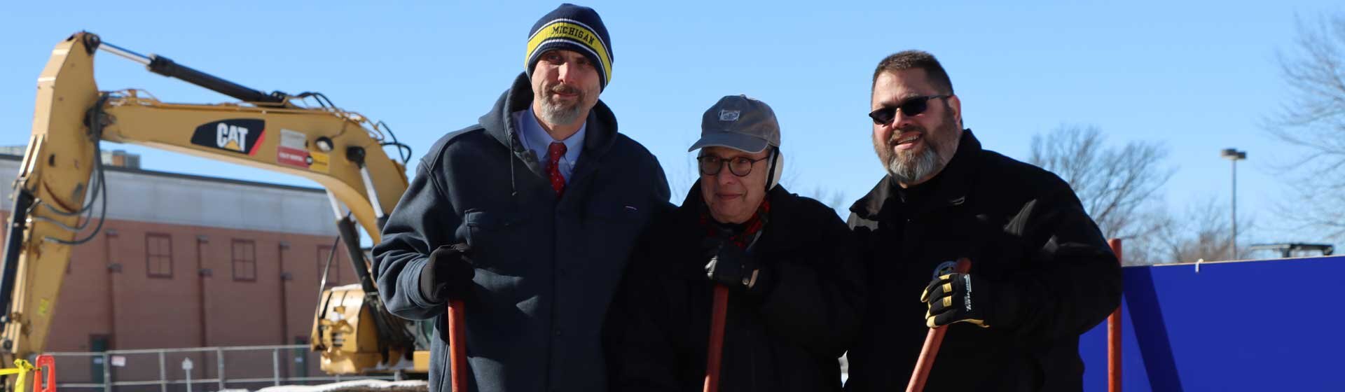 3 men holding shovels with heavy machinery in background