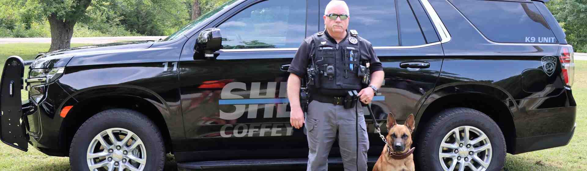 K-9 owner and his brown dog stand in front of their sheriff vehicle.