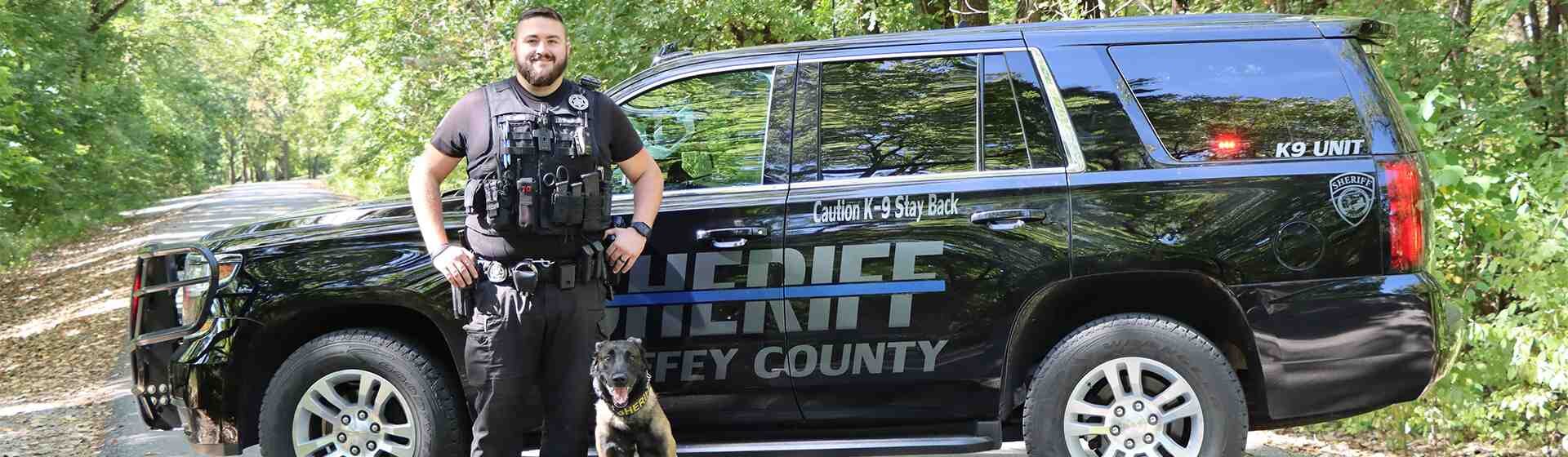K-9 owner and his black dog stand beside their sheriff vehicle.
